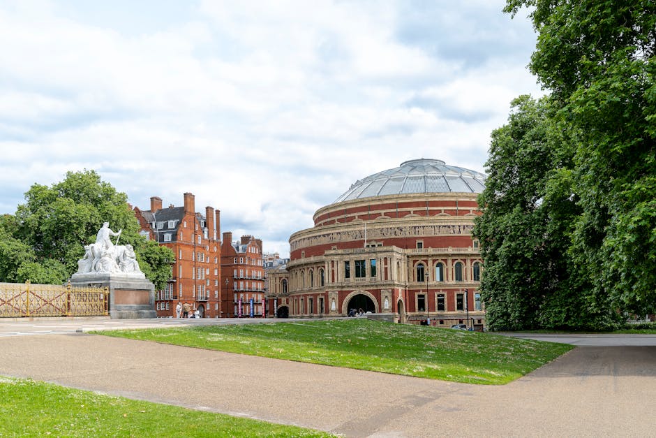 The image depicts a historic multi-storey red brick building with white decorative detailing and multiple bay windows, situated beneath a clear sky. Tall trees with green foliage partially frame the u