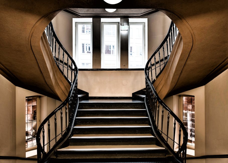 A black metal staircase with wooden steps viewed from the top looking downward, spiraling in a rectangular pattern within a white-walled interior, with a narrow landing at each level. The staircase fe